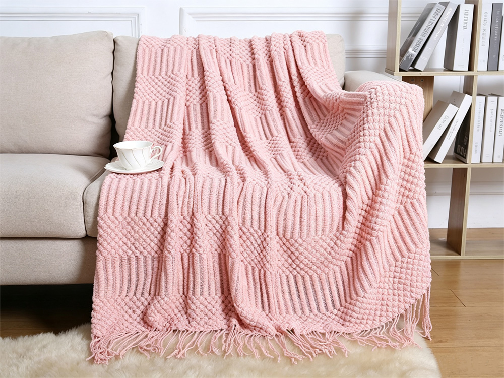 Blush pink bobble knit tassel throw blanket draped over light beige sofa, ceramic teacup, bookshelf background, bright cozy self care living room scene