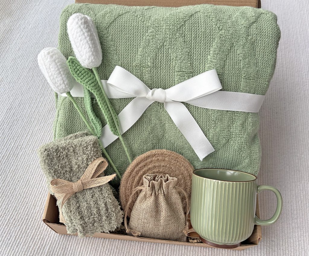 Nestlygift sage green self care gift box, folded textured knit blanket, crocheted tulips, ceramic mug, fuzzy socks and linen sachet flat lay
