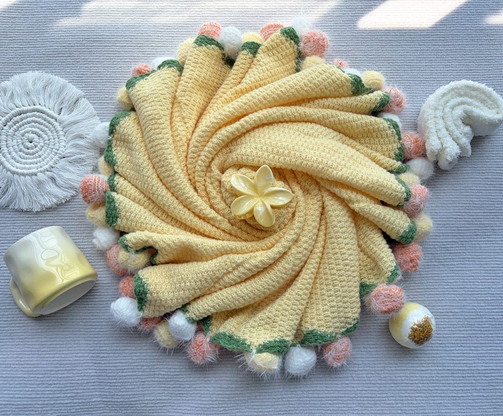 Bright yellow pom pom knit throw blanket arranged in spiral shape, gradient ceramic mug, plush socks, woven coaster, cozy self care flat lay scene