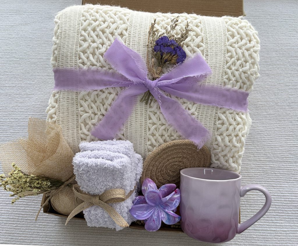 Nestlygift lavender purple self care gift set, thick white knit blanket, dried flowers, gradient ceramic mug and plush socks flat lay